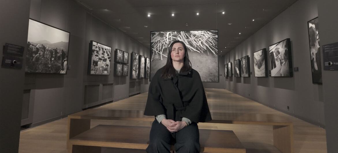 A woman with dark hair sits on a bench in a memorial hall, surrounded by black and white photographs on the walls.
