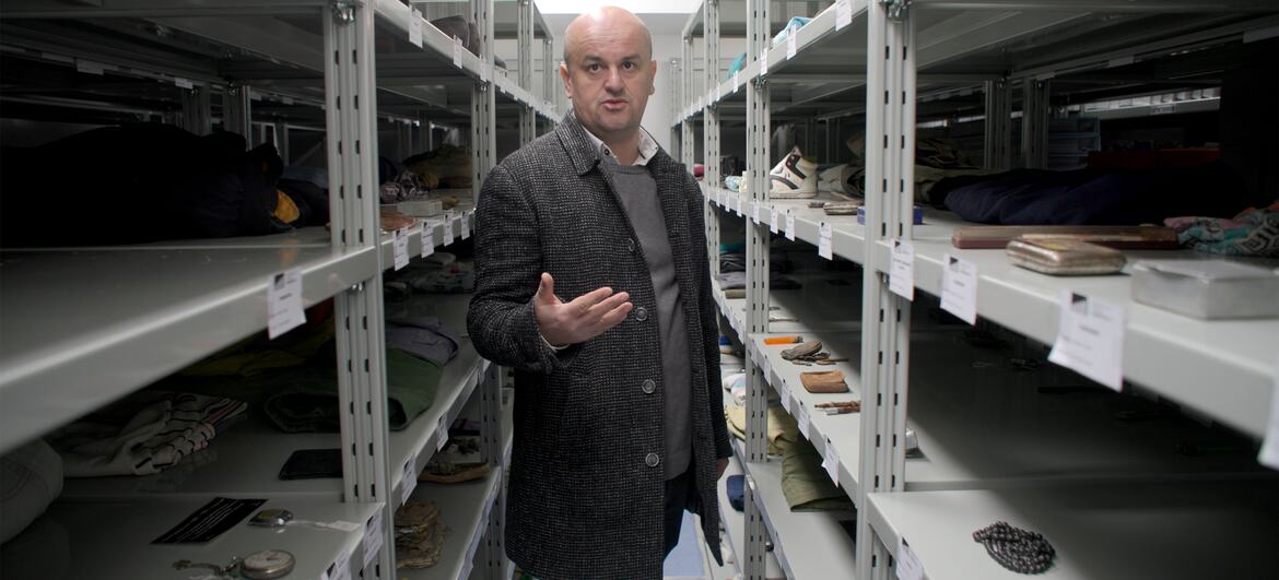 A man in a dark coat stands in a memorial archive, surrounded by shelves containing personal items from the Srebrenica massacre.